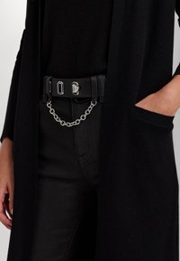 Person som bär svarta byxor och en lång svart cardigan med ena handen i fickan och ett svart bälte med en silverskedja.