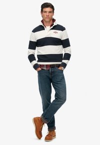Binnenmodel in een marineblauw en wit gestreept polo-shirt, blauwe jeans en bruine laarzen. Opmerkelijke details zijn een rood logo en gelaagd ruitpatroon.