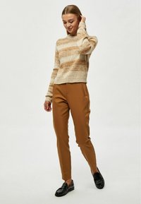 Maglione a righe beige e nocciola lavorato a maglia con scollatura rotonda, abbinato a pantaloni attillati marroni e mocassini neri. Texture morbida e linee pulite.
