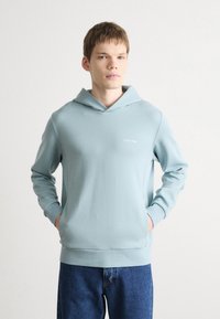 Svetlo modrá mikina s kapucňou a predným vreckom, vyrobená z hladkej látky. Má šnúrku na kapucni a malé biele logo na hrudi.
