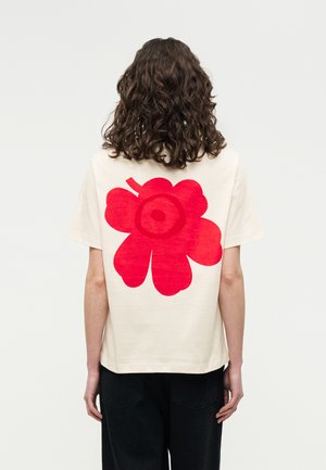 Persona con cabello rizado que lleva una camisa blanca con un gran diseño abstracto de una flor roja en la parte trasera, combinada con pantalones negros.