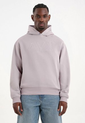 Uomo con felpa con cappuccio viola chiaro con logo "AVX" e jeans blu, in piedi contro uno sfondo bianco uniforme.