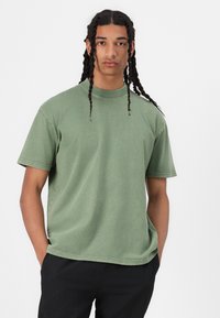 Grüne Baumwoll-T-Shirt mit kurzen Ärmeln, Rundhalsausschnitt und lockerer Passform. Dezent strukturierte Textur und Steppnähte entlang der Säume.