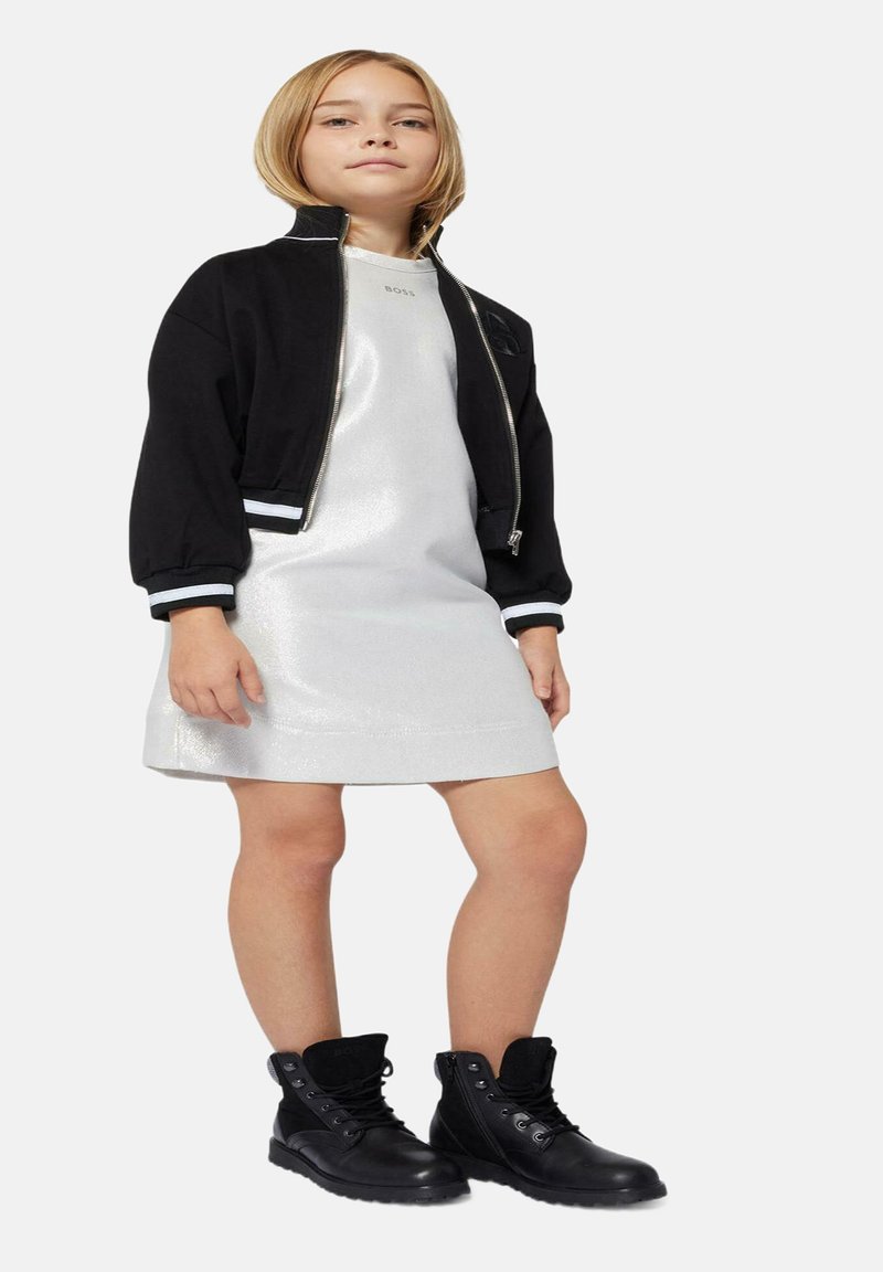 Veste bomber noire avec des accents blancs sur une robe argentée réfléchissante. Bottes noires à lacets avec un détail texturé.