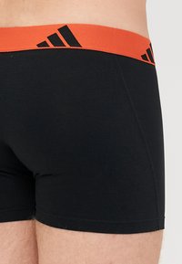 Črne bokserice z oranžnim elastičnim pasom z črnim Adidasovim logotipom. Gladka tekstura, prileganje in dolžina do sredine stegna.