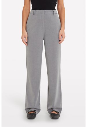 Femme portant un pantalon gris taille haute à jambes larges, associé à des ballerines noires et un haut noir sans manches, debout sur un fond blanc.