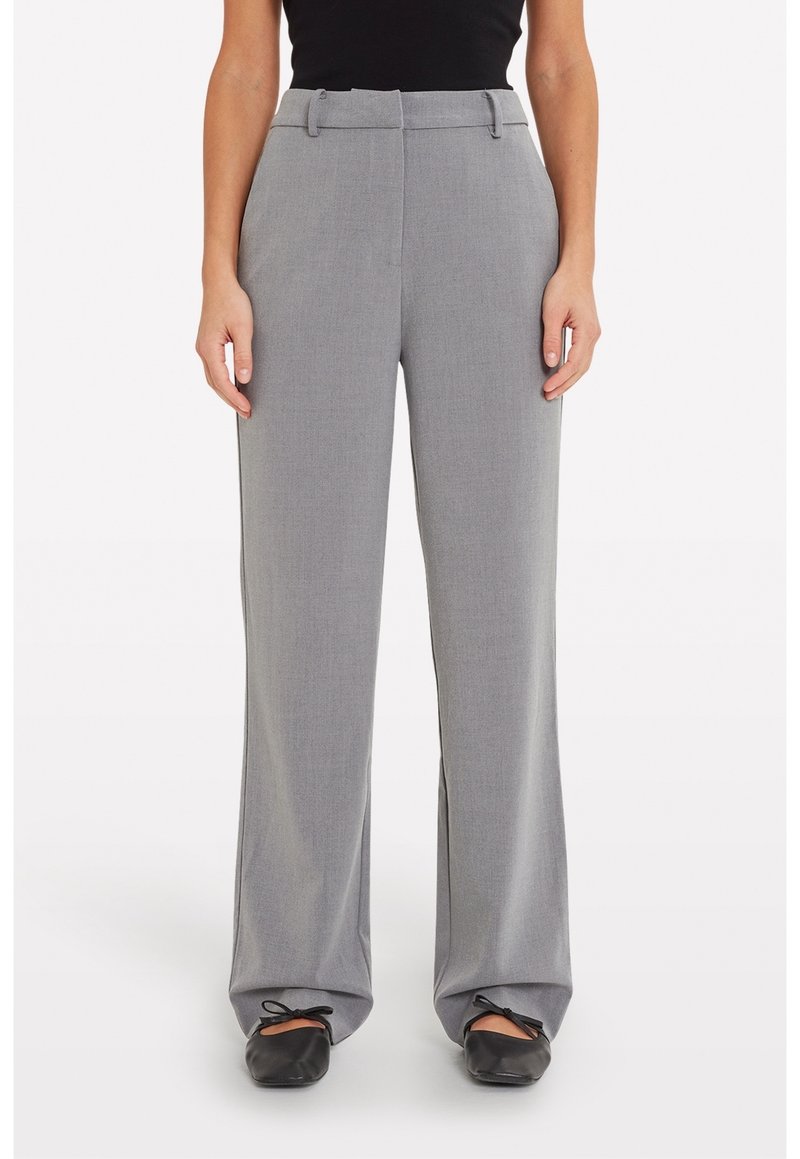 Femme portant un pantalon gris taille haute à jambes larges, associé à des ballerines noires et un haut noir sans manches, debout sur un fond blanc.
