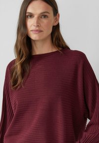 Bordowy top w paski z długim rękawem, szerokim dekoltem i luźnym krojem. Materiał wydaje się miękki, o delikatnej fakturze, swobodnie układa się na ciele.