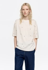 Kremowa koszulka z krótkim rękawem o okrągłym dekolcie, ozdobiona małymi czarnymi detalem, w zestawie z ciemnoniebieskimi spodniami z szerokimi nogawkami.