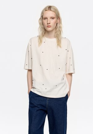 Camiseta corta de manga corta con cuello redondo, adornada con pequeños detalles negros, combinada con pantalones de pierna ancha de color azul oscuro.