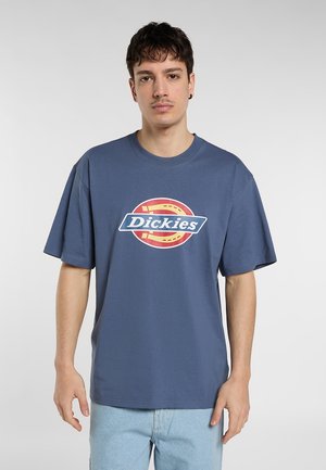 Joven con cabello oscuro corto que lleva una camiseta azul holgada con el logo de Dickies y vaqueros azul claro, de pie contra un fondo liso.