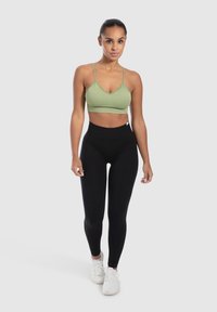 Sujeción deportiva verde con canalé y tirantes finos, combinada con leggins negros de cintura alta. Zapatillas blancas y un fondo neutro.