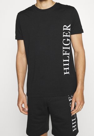 Homme portant un ensemble noir composé d’un t-shirt à manches courtes et d’un short avec un texte vertical blanc "HILFIGER" sur le côté gauche du t-shirt et du short.