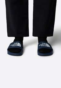Modre sanjske sandale s širokim trakcem, na katerem je logo "The North Face". Nosi se z črnimi nogavicami in v kombinaciji s temnimi hlačami.