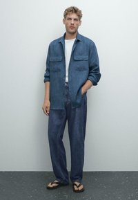 Uomo che indossa una camicia blu a bottoni sopra una maglietta bianca, jeans scuri larghi e infradito neri, in piedi contro un muro grigio chiaro.