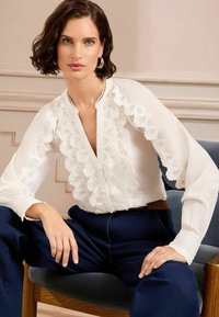 Blusa bianca a maniche lunghe con bordo in pizzo scallop, chiusura a bottoni e vestibilità comoda, abbinata a pantaloni blu navy e collocata su uno sfondo neutro.