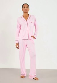 Femme debout pieds nus portant un ensemble de pyjama à manches longues boutonnières rose clair avec des bordures rouges au col, aux poignets, à la poche et aux ourlets des pantalons.