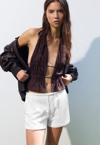 Hnedý textúrovaný crop top so zhromaždenými detailmi, kombinovaný s bielymi šortkami na sťahovanie. Tmavo hnedá oversized bunda, model pózujúci na svetlom pozadí.