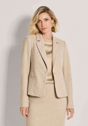 Frau mit kurzen blonden Haaren, die einen beigen Blazer, einen passenden Rock und einen satinierten beigen Top trägt, vor einem neutralen Hintergrund stehend.