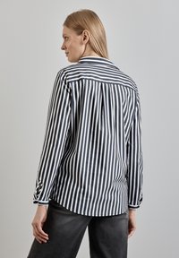 Camicia a righe bianche e nere con colletto abbottonato, maniche lunghe e orlo curvo. Il tessuto sembra liscio con una leggera lucentezza.