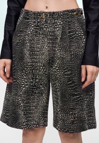 Shorts texturés noirs et beiges avec un motif abstrait de reptile, portés avec un haut noir à manches longues, laissant apparaître le ventre et les mains.