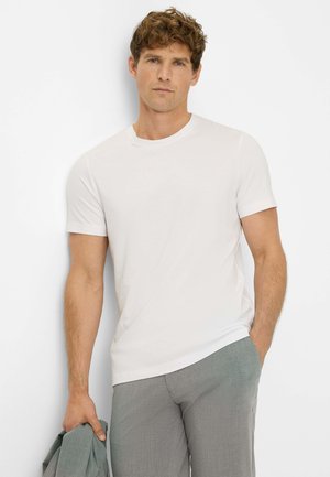 Weißes T-Shirt mit kurzen Ärmeln und Rundhalsausschnitt, aus glattem Baumwollstoff, figurbetontes Design, kombiniert mit hellgrauen Hosen, Model hält eine Jacke.