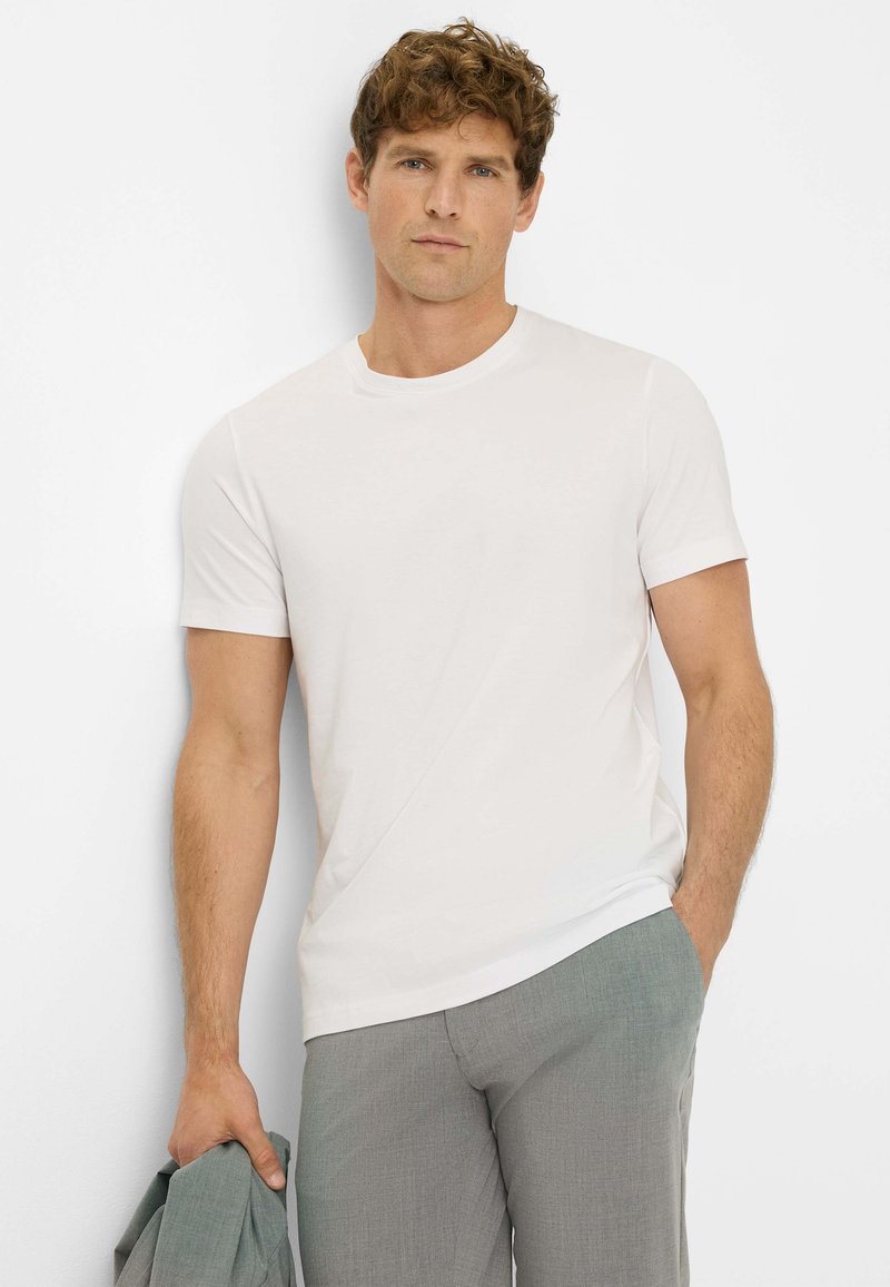 T-shirt bianca a maniche corte con scollatura rotonda, in morbido cotone, design aderente, abbinata a pantaloni grigi chiari, modello che tiene in mano una giacca.