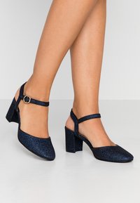 Mörkblå, glittriga slingback-hälar med rund tå, blockklack och ett smalt ankelsnöre med en cirkulär metallspänne.