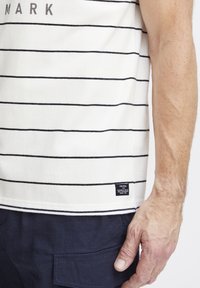 Dryžuota švelniame ir tamsiai mėlyname tanko marškinėlyje; minkšta medvilnės medžiaga. Yra mažas logotipo ženklelis prie apačios ir laisvas kirpimas, pabrėžiantis ranką.