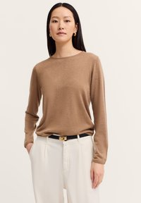 Frau mit langen glatten Haaren, die goldene Creolen trägt, einen hellbraunen Langarmoberteil, weiße hoch geschnittene Hosen und einen schwarzen Gürtel.