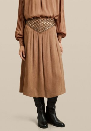 Vestido marrón con plisados y un acabado texturizado, que presenta una cintura adornada y mangas largas, combinado con botas negras hasta la rodilla.