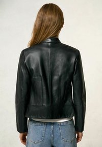 Veste en cuir noire à coupe ajustée, avec col, coutures verticales et ourlet légèrement raccourci, portée sur un jean bleu clair.