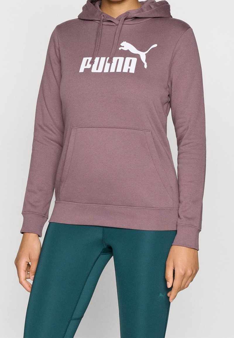 Personne portant un sweat à capuche mauve Puma avec poche avant et des leggings bleu canard foncé.