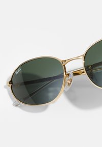 Gafas de sol aviador doradas con lentes de un verde oscuro, con un marco de metal y almohadillas nasales transparentes. Logo de Ray-Ban visible en la lente.