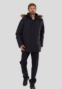 Man in een zwarte geïsoleerde parka met een met bont gevoerde capuchon, zwarte broek en zwarte schoenen, staande tegen een effen lichtgrijze achtergrond.
