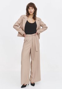 Beżowy garnitur o dopasowanym kroju, z dopasowaną marynarką i szerokimi spodniami, z zawiązanym w pasie detalem. Czarna bluzka i szpilki dopełniają stylizację.