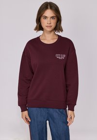 Sweatshirt bordeaux avec un col rond et des poignets côtelés. Présente "CITY CLUB" brodé en blanc sur la poitrine gauche. Conception à coupe décontractée.