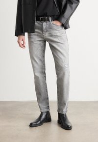 Jeans gri din denim cu o croială slim, prezentând detalii uzate și o nuanță discretă de estompare, asortați cu cizme negre din piele până la gleznă.