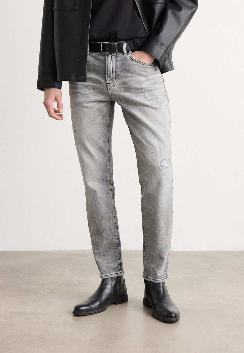Jeans gri din denim cu o croială slim, prezentând detalii uzate și o nuanță discretă de estompare, asortați cu cizme negre din piele până la gleznă.
