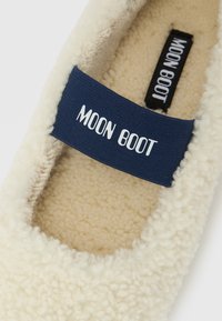 Pantufla de forro polar color crema con una correa azul marino etiquetada "MOON BOOT" en la parte superior y en la suela interior.