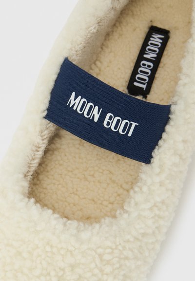 Chausson en polaire couleur crème avec une bride bleu marine portant l'inscription « MOON BOOT » sur le dessus et à l'intérieur de la semelle.