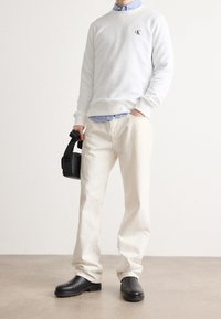Sudadera blanca con logo negro, superpuesta a una camisa de rayas azul. Pantalones beige claros y botas de cuero negras. Bolsa negra en la mano.