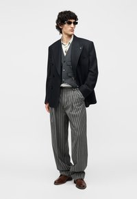 Άνδρας που φοράει σκούρο σακάκι, γκρι γιλέκο, ριγέ παντελόνι με φαρδιά μπατζάκια, καφέ παπούτσια, γυαλιά ηλίου και μενταγιόν, στέκεται με το ένα χέρι στην τσέπη.