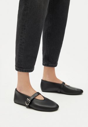 Scarpe basse in pelle nera con punta arrotondata e design traforato, caratterizzate da una fascia con fibbia sul collo del piede. Indossate con jeans scuri.