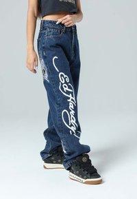 Pantalones de mezclilla azul con un corte holgado, con texto gráfico y diseño en blanco en la pierna izquierda. Combinados con zapatillas negras con acentos decorativos.