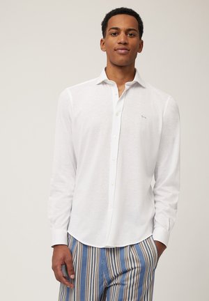 Camisa blanca de manga larga en tejido ligero con cuello abotonado, con un discreto bordado de logo azul en el pecho.