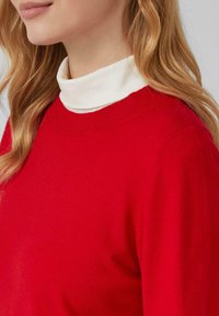 Czerwony sweter z okrągłym dekoltem, wykonany z miękkiego materiału, nałożony na biały golf. Gładka faktura, brak widocznych wzorów lub elementów metalowych.