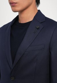 Sötétkék blézer texturált felülettel, éles hajtókákkal és fekete virágmintás tűződíszítéssel a galléron. Sötét pulóver felett viselve.