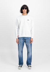 Mann med krøllete hår iført svarte solbriller, hvit sweatshirts med logo, blå jeans og svarte sneakers mot en ensfarget hvit bakgrunn.