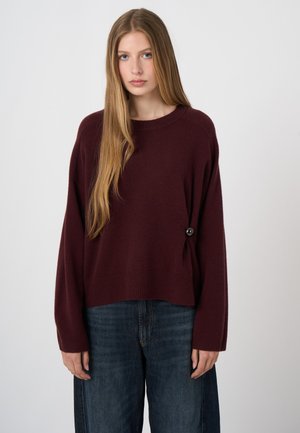 Vrouw met lang blond haar draagt een losse bordeauxrode trui met een zijknop en brede donkerblauwe spijkerbroek tegen een effen achtergrond.